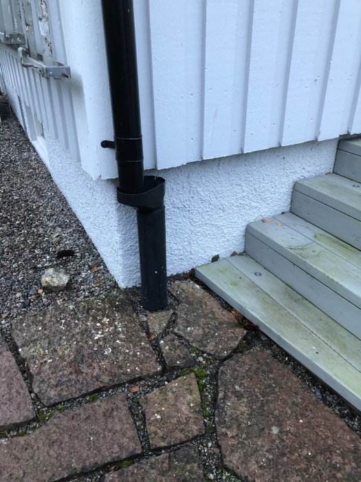 Hörnet av ett hus med vit korrugerad vägg och en svart stuprör nedför till marken intill stenplattor och trätrapp.