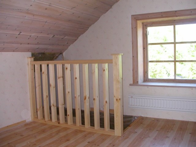 Trægelænder på anden etage ved siden af et skråloft og et vindue. Væggene er hvidmalede, og der er lyst trægulv.