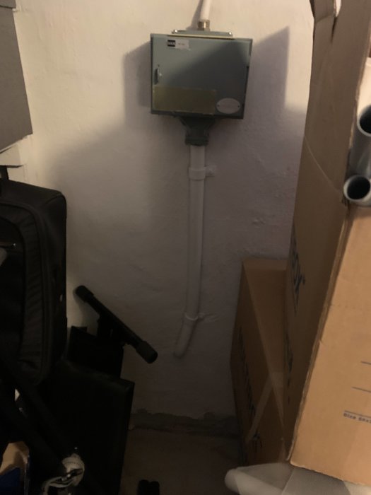 Elservis med grå skåp och rörledningar på en vägg i en källare med kartonger och föremål i förgrunden.