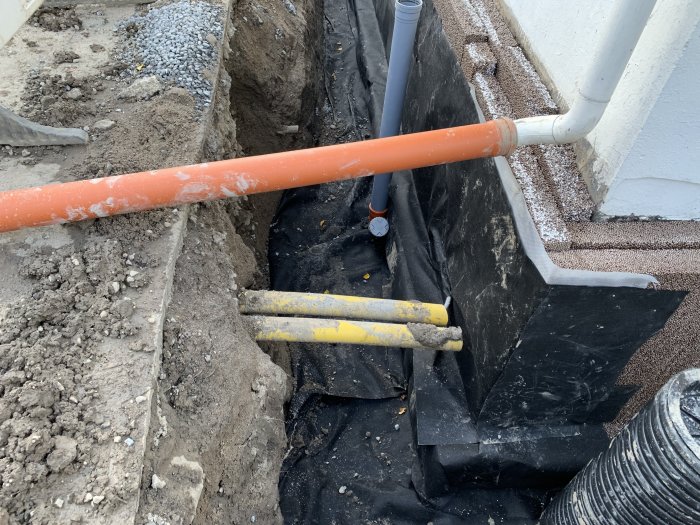 Grävd dräneringsränna vid husvägg med synlig anslutningspunkt och isolering.