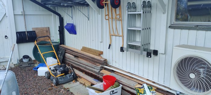 Byggmaterial staplat mot garagevägg under tak med trädgårdsredskap och luftvärmepump.