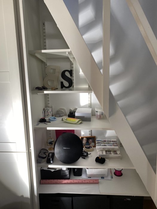 Platsbyggda hyllplan under en trappa upp till en loftsäng, öppna och fyllda med diverse föremål.