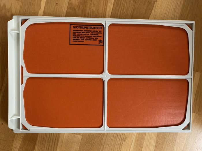 Vit plastback med fyra orangea fack och skötselinstruktioner på en träyta.