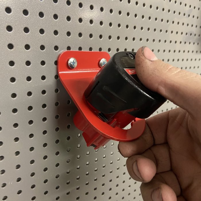 Hand håller en nyligen pulverlackerad röd batterihållare monterad på en perforerad verktygsvägg.
