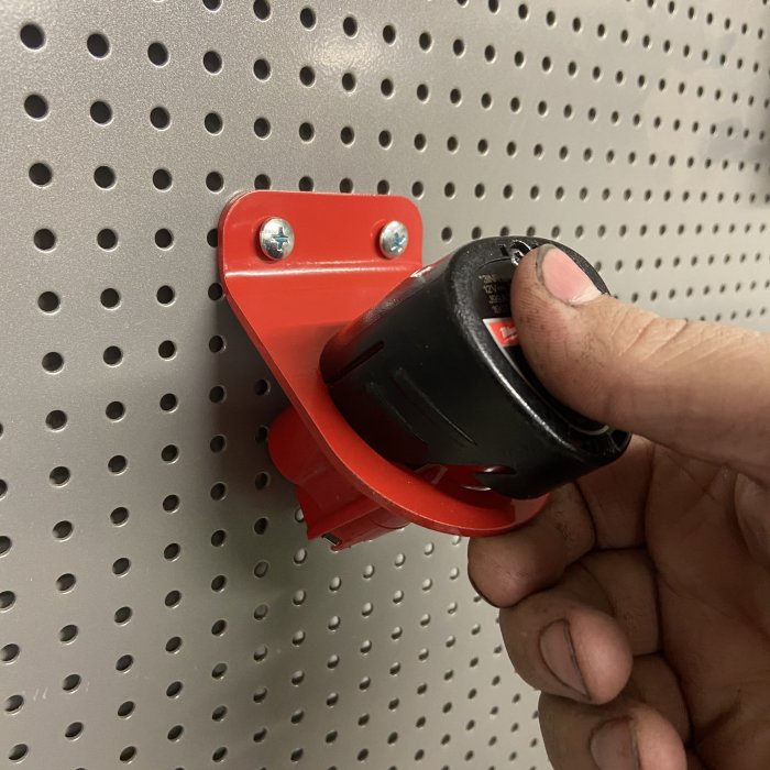 Hand som placerar en batterihållare på en röd, bockad och pulverlackerad metallhållare fäst på en perforerad vägg.