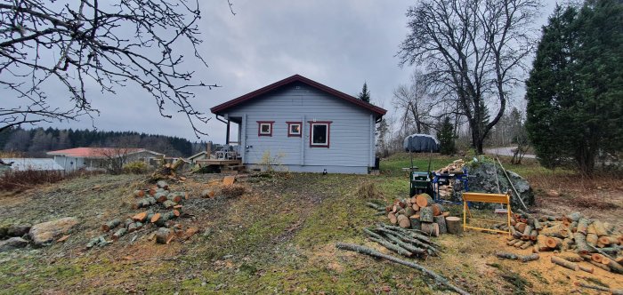 En grävare nära en stapel av nysågade trästammar framför ett hus med plintgrund under byggnation.