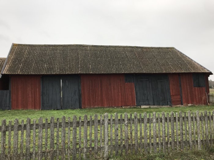 Röd lada med slitet tak och väggar som bågnar, framförallt på ena sidan, staket i förgrunden.