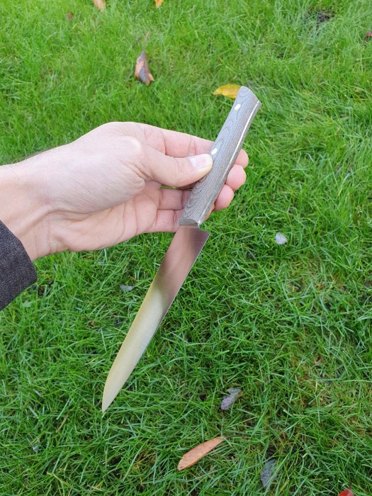 Hand håller en liten handgjord kökskniv med trähandtag mot gräsbackgrund.