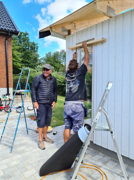 Två män monterar en trädetalj på en vit byggnad med verktyg och stege framför.