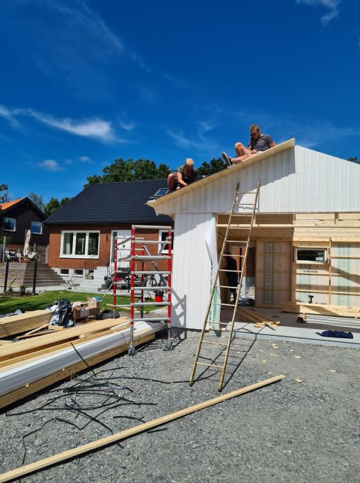 Två personer sitter på taket till en halvfärdig träbyggnad under konstruktion mot blå himmel.