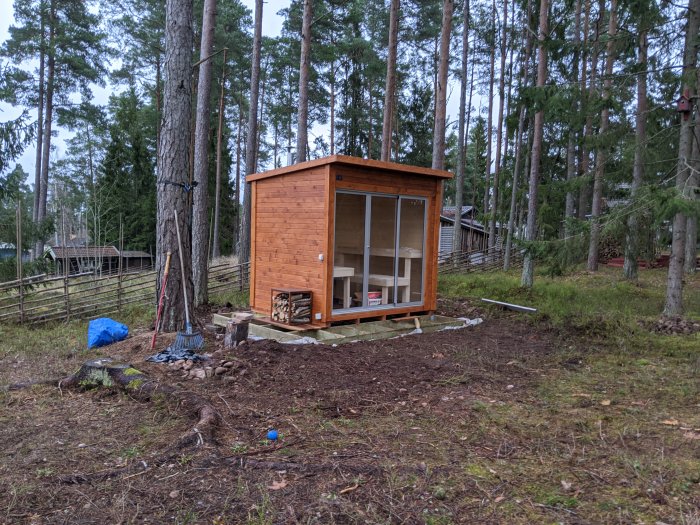 Nyinstallerad bastustuga med klarglas på en altan i en skogstomt, väntar på trall.