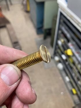 Hand håller en nysvarvad försänkt mässingsskruv anpassad för att spännas med fingrarna i en verkstadsmiljö.