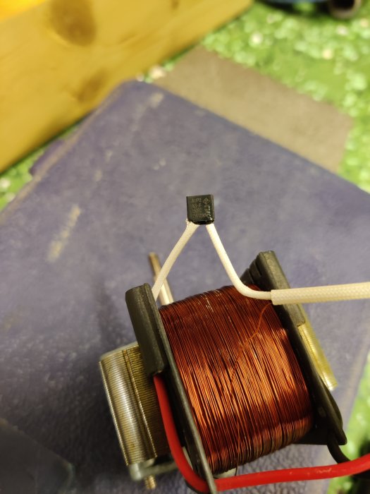 En elektronisk komponent hålls med pincett vid en kopparlindad spole, antydan till skada på komponenten.