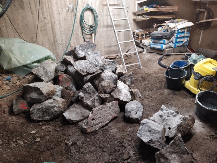 En hög med stora bergstenar på en byggarbetsplats inuti en byggnad, med en stege och arbetsmaterial i bakgrunden.