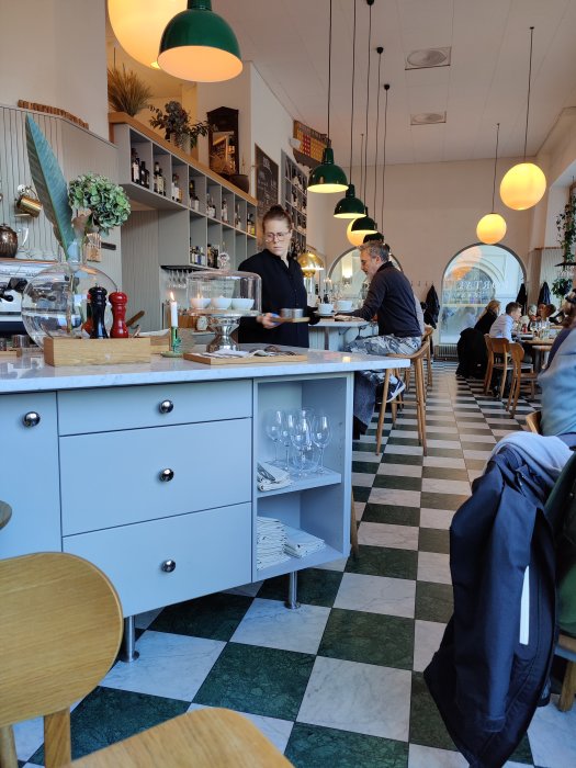 Interiör av en restaurang med serveringspersonal, matgäster, bardisk och hängande lampor.