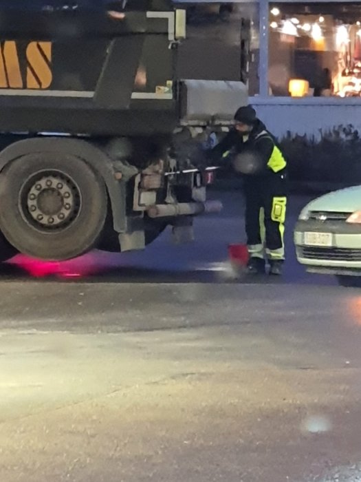 En person i reflexkläder tvättar en lastbilshjul med en hink och borste på en bensinmack om kvällen.