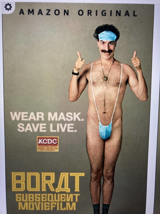 Man i Borat-kostym och munskydd ger tummen upp framför kampanjtext för maskanvändning.