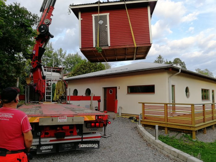 Ett hus lyfts av en mobilkran medan en person iakttar; reflektion över det krävande byggprojektet.