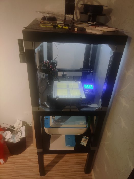 Ender 3 3D-skrivare i anpassat skåp med LED-belysning och filamentrullar på översta hyllan.