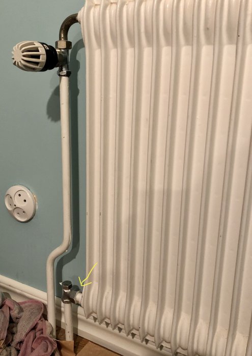 Radiator med termostat och returventil markerad med en pil, handduk och eluttag i bakgrunden.