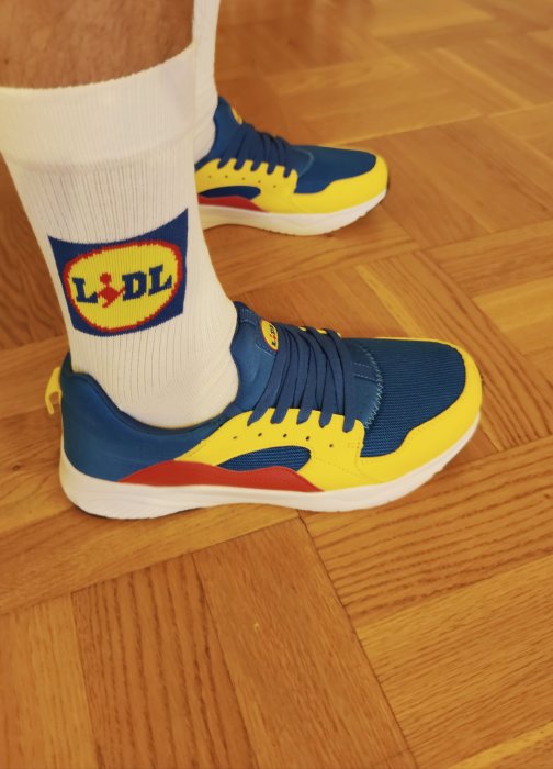 Person som står på trägolv med sportiga blå och gula sneakers samt vita strumpor med livsmedelskedjans logga.