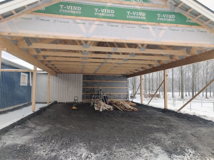 Halvfärdigbyggd carport av trä med takstolar och vindskydd, högra sidan delvis panelad, vänster öppen.