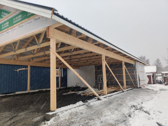 Halvfärdig träkonstruktion av en carport med takstolar synlig och en delvis täckt sidovägg, i vintersnö.