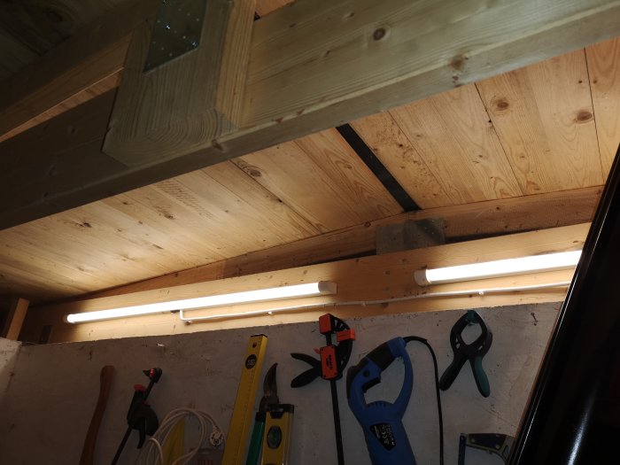Bastupanel på innertak med synliga takbjälkar och lysrör, verktyg på arbetsbänk nedanför.