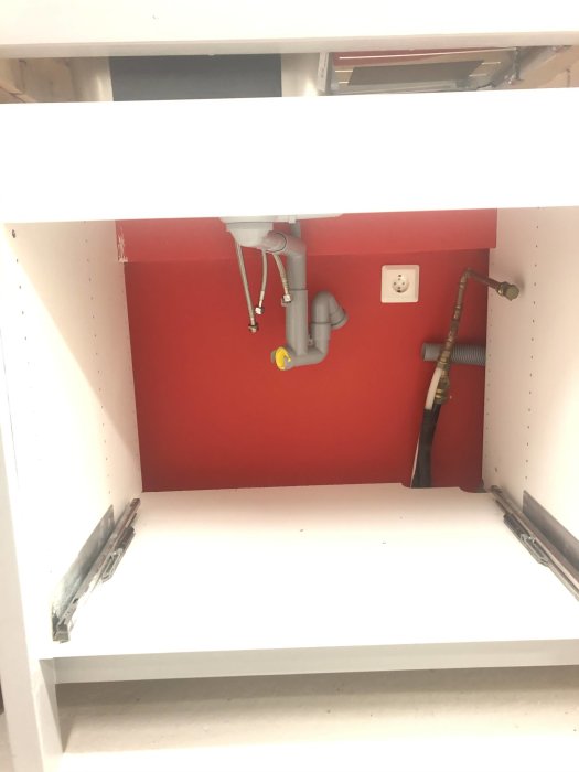 Utrymme under diskbänk med synliga avloppsrör, vattenanslutningar och eluttag, redo för installation av varmvattenberedare.