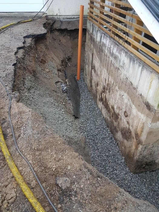 Uppgrävd dränering vid husgrund med stuprör, grus och synlig isolering utan avrinningssystem.