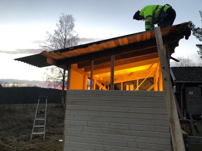 Arbetare installerar takplåt på en byggnad med träytterpanel och belyst innertak vid skymning.