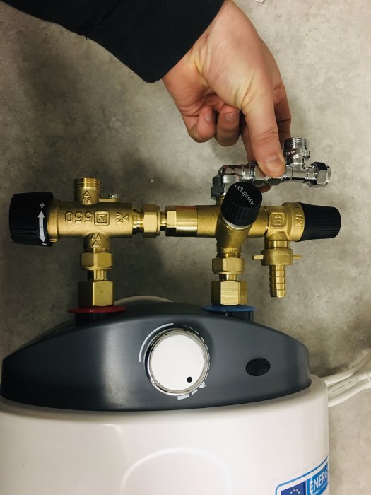 En hand som justerar en installerad blandarventil ovanpå en vit varmvattenberedare, med olika mässingskopplingar och rör.