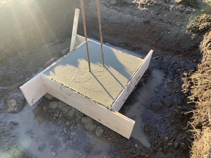 Nyutgjuten betongfundament med armeringsjärn och formar av trä i en grävd grop.