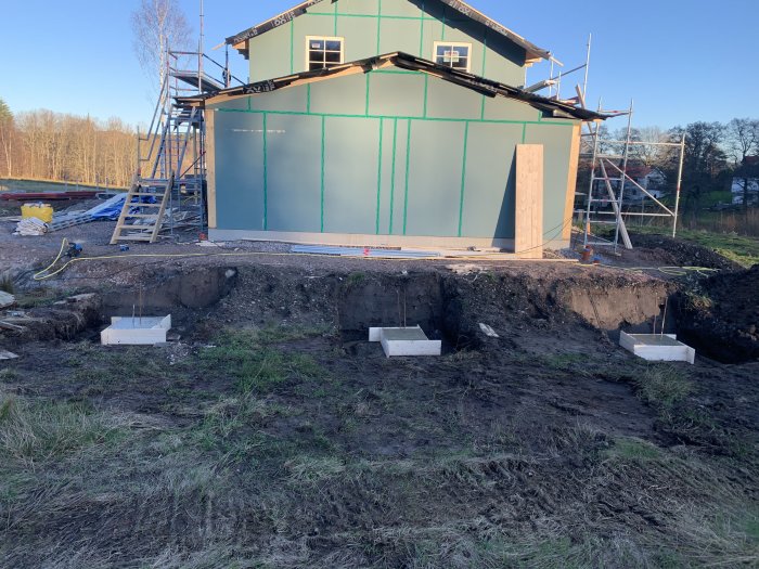 Pågående byggnation med gjutna fundamentklumpar för carport och ett halvfärdigt hus med armeringsjärn och skyddad plåtremsor runt fönster.