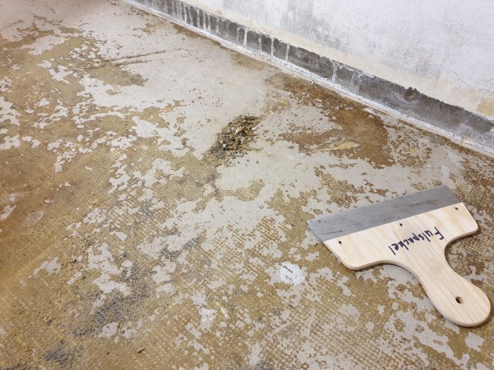 Betonngolv med kvarvarande gulaktigt sprött lim och rester från mattans undersida, spatel ligger på golvet.