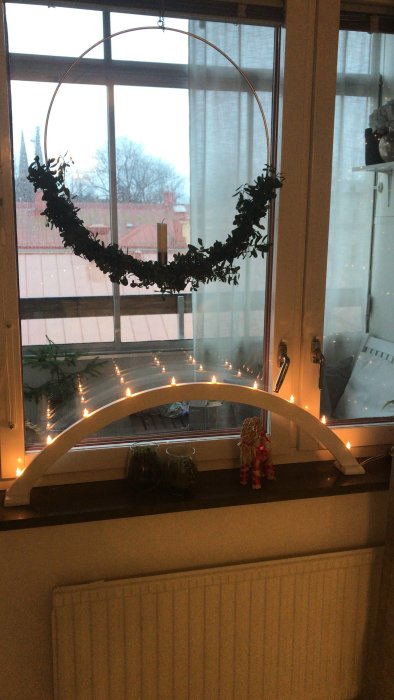 Fönster med juldekorationer, ljusslinga på båge och krans, mörkt ute.