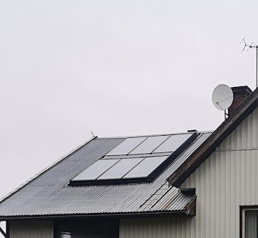 Timmerhus med solfångare på taket, parabolantenn och snedtak med regnig himmel i bakgrunden.