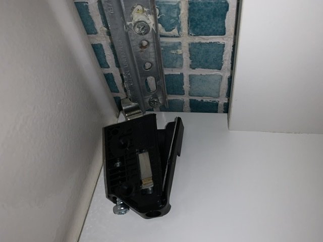Svart justerbart fäste för badrumskommod lossnat från väggskena, med delvis synlig text 1975N UNI 10768, i hörn med kakel.