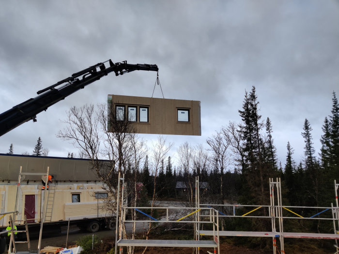 Lyftkran som höjer en stor väggsektion med fönster för montage i ett bygge, mot skogsbakgrund och molnhimmel.