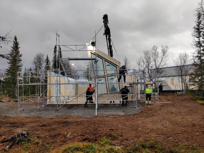 Byggarbetsplats med arbetare som monterar ytterväggar på husstomme omgiven av byggställningar.