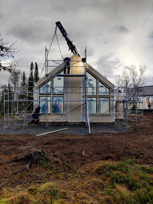 Hus under konstruktion med ytterväggar på plats, byggnadsställningar runt om, och en kran som lyfter material.