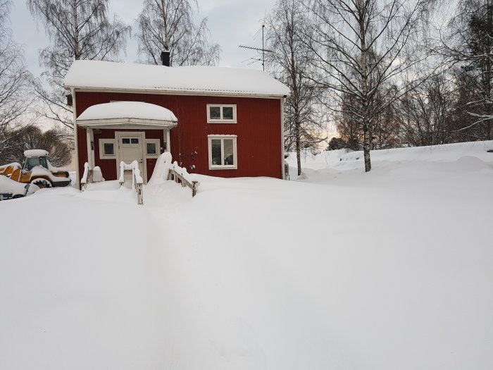 Röd gårdshus fasad täckt av snö med spår efter snöröjning och snötyngda träd i bakgrunden.