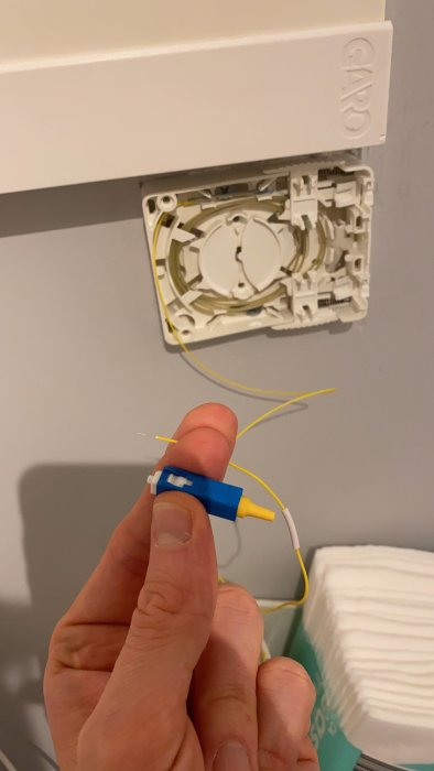 En person håller en blå fiberoptisk kontakt med en avsliten gul fiberkabel framför en öppen fast dosa för fiberanslutning på väggen.