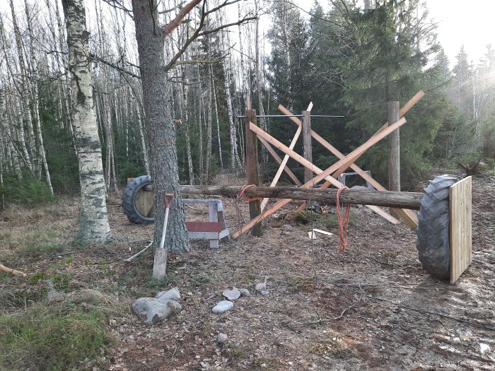 Delvis konstruerad gungbräda med sidlånge trästolpar och genomfört rör, omgiven av träd och däck i ett skogsområde.