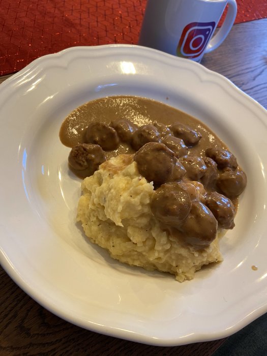 Hemgjort potatismos med köttbullar och brunsås serverat på en vit tallrik, vid en röd duk.