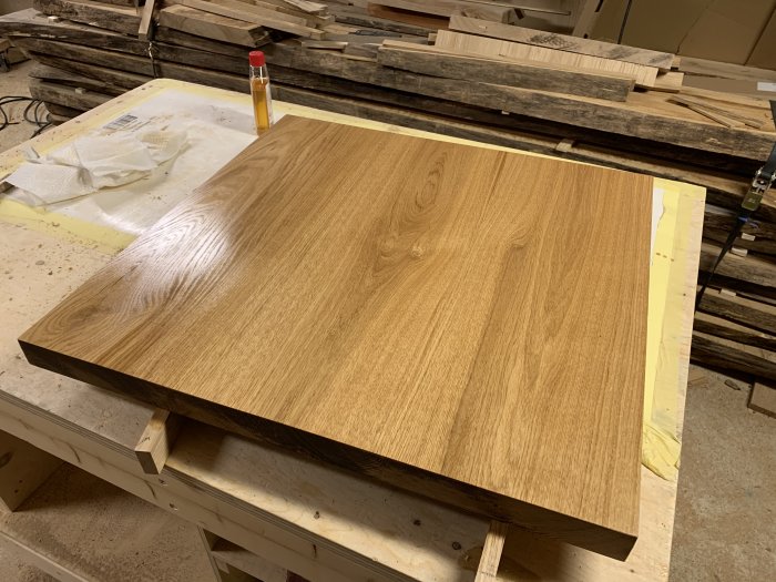 Träbänkskiva med slutfinish i ett snickarverkstad, omgiven av verktyg och obehuggna plankor.