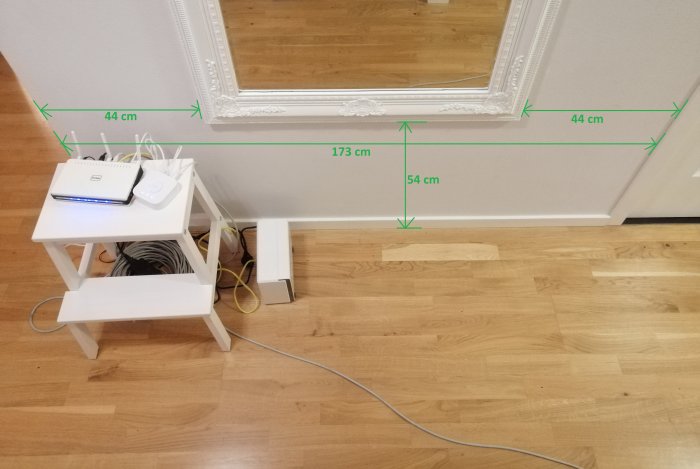 En hall med elektronik på en vit liten hylla och kablar på golvet. Gröna måttangivelser visar den tänkta möbelns placering och storlek.