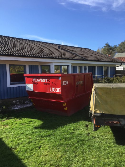 En röd mindre container framför ett blått hus under en rivningsprocess.