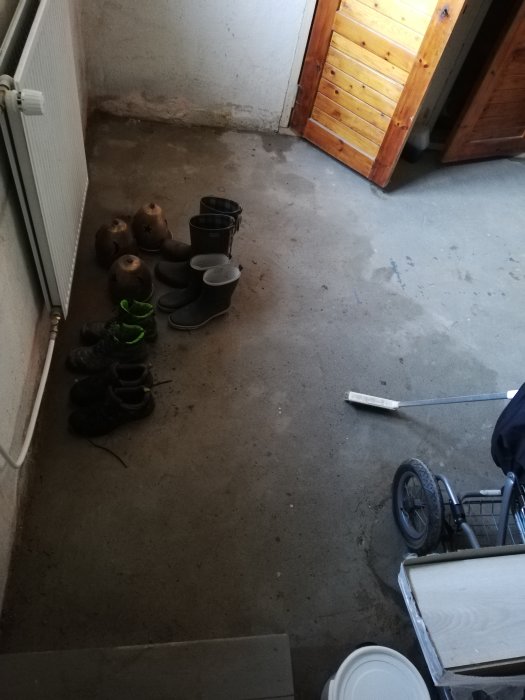 Källare med synliga fuktspår på betonggolv och väggar, stövlar och föremål på golvet.