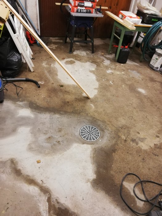 Källargolv med fuktfläckar och avloppsbrunn, verktygsbänk och byggmaterial i bakgrunden.